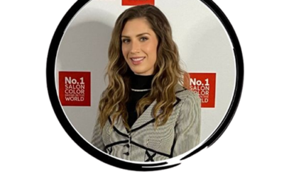 Smiling woman with wavy light brown hair posing for a photo at a Salon Color World event with No.1 Salon Color World signs behind her.