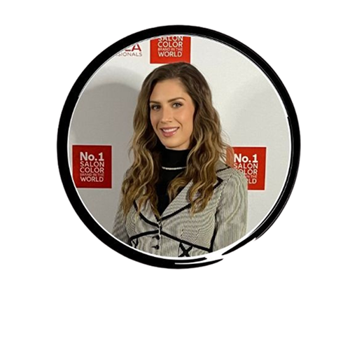 Smiling woman with wavy light brown hair posing for a photo at a Salon Color World event with No.1 Salon Color World signs behind her.